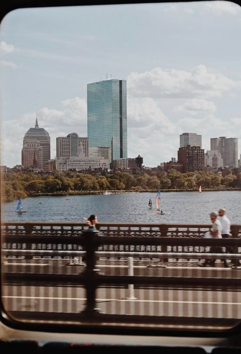 Boston skyline viewed through a train window, with sailboats on the Charles River and the John Hancock Tower in the background